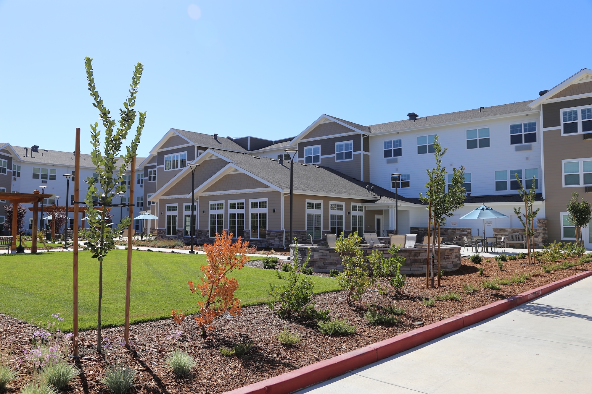 A modern residential building with three stories, large windows, and a neutral color palette. The landscaped yard features young trees, shrubs, and a patio area with umbrellas. Clear blue sky in the background on a sunny day.