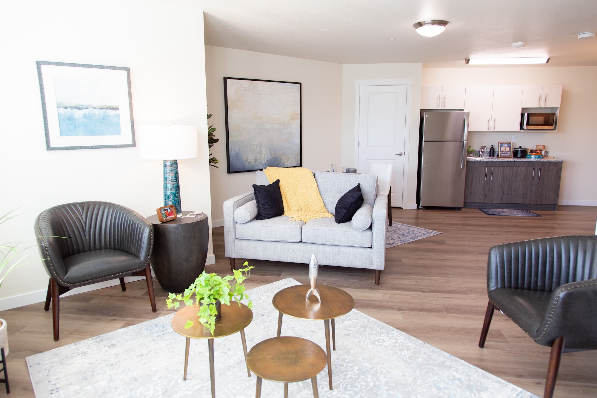 A modern living room with a gray sofa, yellow throw, and two dark armchairs. A round coffee table set with plants in the center, a floor lamp, and artwork decorate the light-colored walls. The kitchen area can be seen in the background.