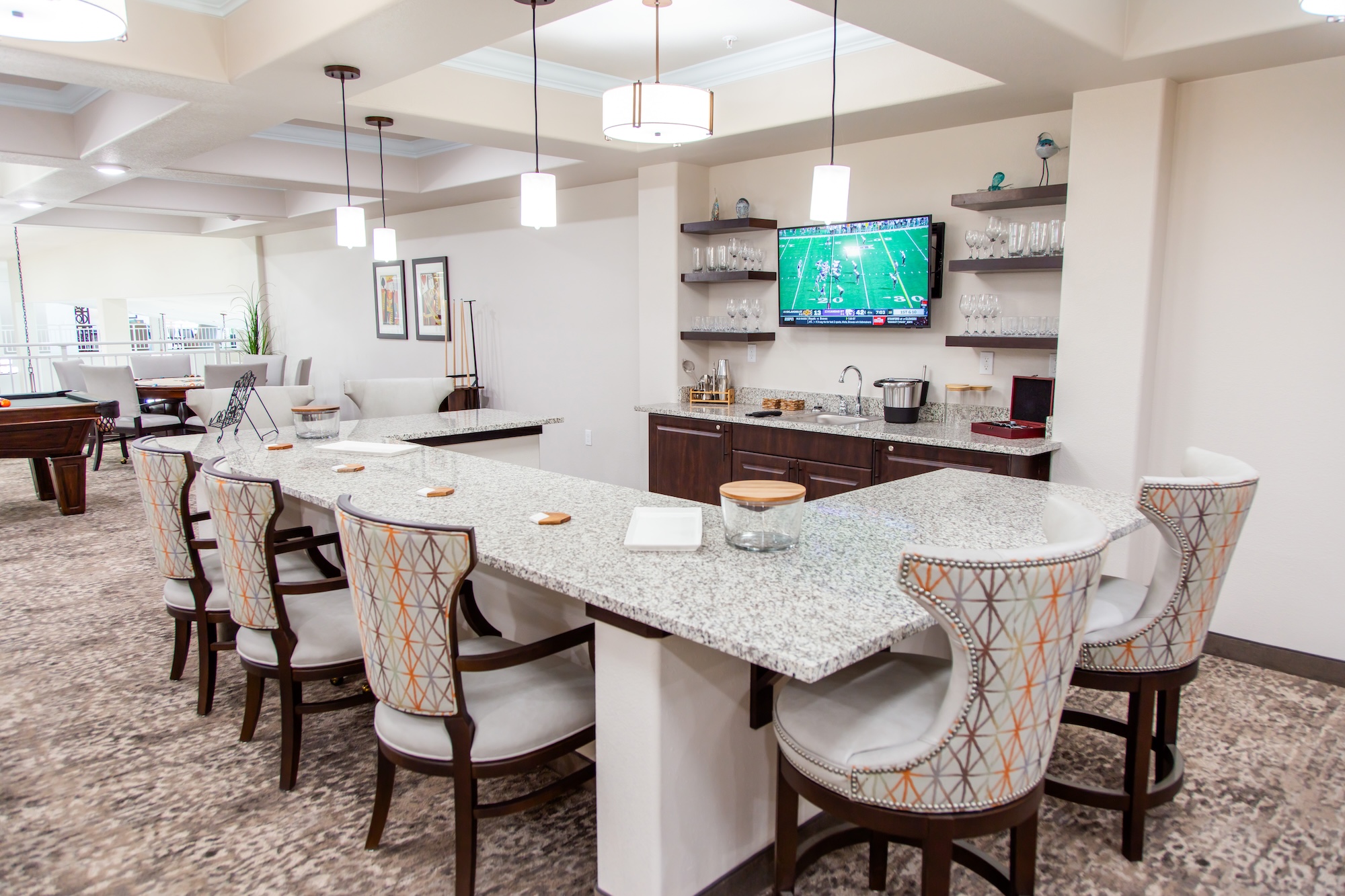 A modern kitchen and bar area featuring a long granite countertop with high-backed, patterned chairs. The walls are decorated with shelves and framed art. A flat-screen TV displays a sports game. The space is well-lit and carpeted.