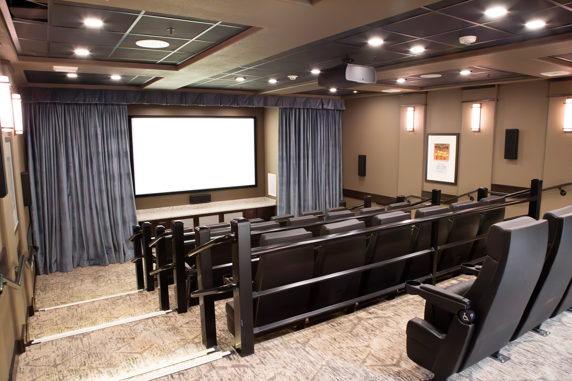 A small, modern movie theater with rows of black leather seats facing a large screen. The room has gray walls, blue curtains, and soft ceiling lights, creating a cozy ambiance. Framed posters are displayed on the walls.