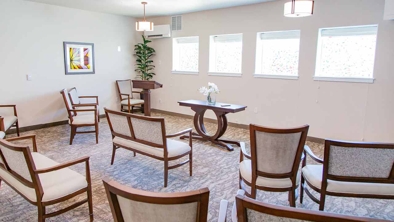 A room with a podium, several upholstered chairs, and a wooden table with flowers arranged in a semicircle. The walls have small windows and a framed artwork. The floor is carpeted, and a potted plant is in the corner. Lighting fixtures hang from the ceiling.