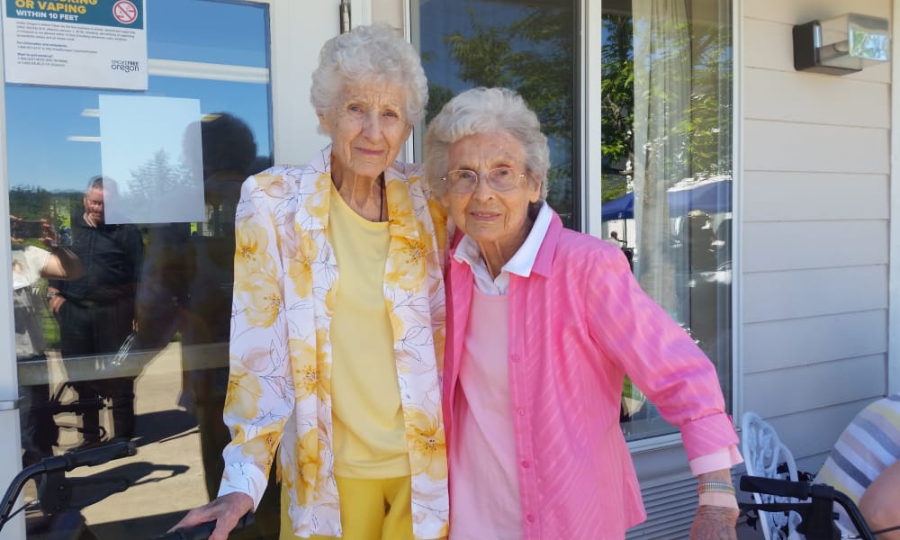 Two elderly women standing close together outside a building with large windows, both smiling. One wears a yellow floral jacket over a yellow top, and the other wears a pink and white striped jacket over a white top. A 
