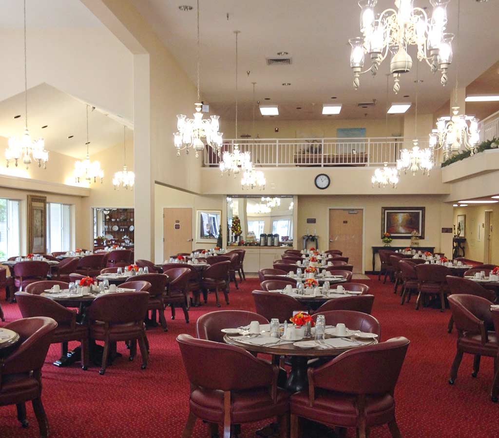 A spacious dining area with red carpet and circular tables, each set with white tableware and red flower centerpieces. Plush red chairs surround the tables, and chandeliers hang from the high ceiling. There is a mezzanine level and a clock on the far wall.