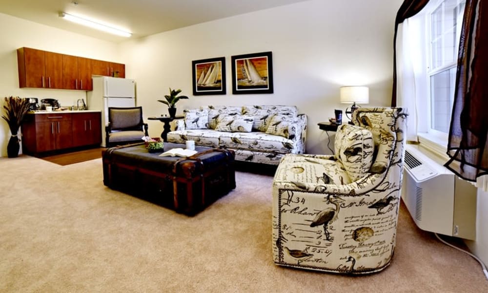 A cozy living room features patterned furniture including a couch and an armchair, a dark wooden coffee table with storage, and framed artwork on the wall. An open kitchen with wooden cabinets, a refrigerator, and a range microwave is seen in the background.