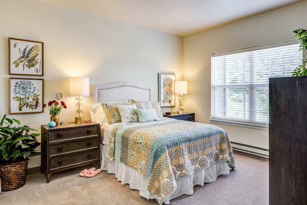 A cozy bedroom with a queen-sized bed adorned with a blue and yellow patchwork quilt and multiple pillows. Two bedside tables with lamps and floral artwork on the walls flank the bed. A large window with blinds lets in natural light, and a plant sits in the corner.
