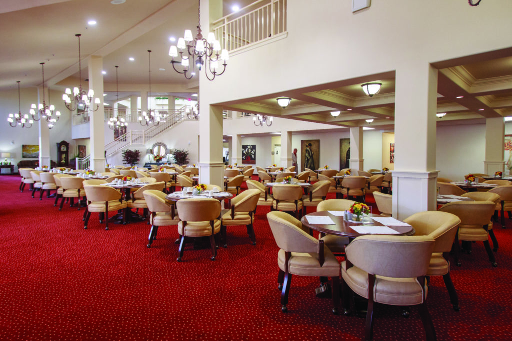 A spacious dining hall with red carpet, numerous round tables, and beige cushioned chairs. Chandeliers hang from the ceiling, and a staircase is visible in the background. Tables are set with tableware and flowers, creating an inviting atmosphere.