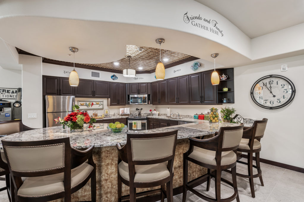 A spacious, modern kitchen with dark wooden cabinets, stainless steel appliances, and a large granite island. Stylish barstools surround the island. Decorative pendant lights hang from the ceiling, and a large clock and wall art add a cozy touch to the room.