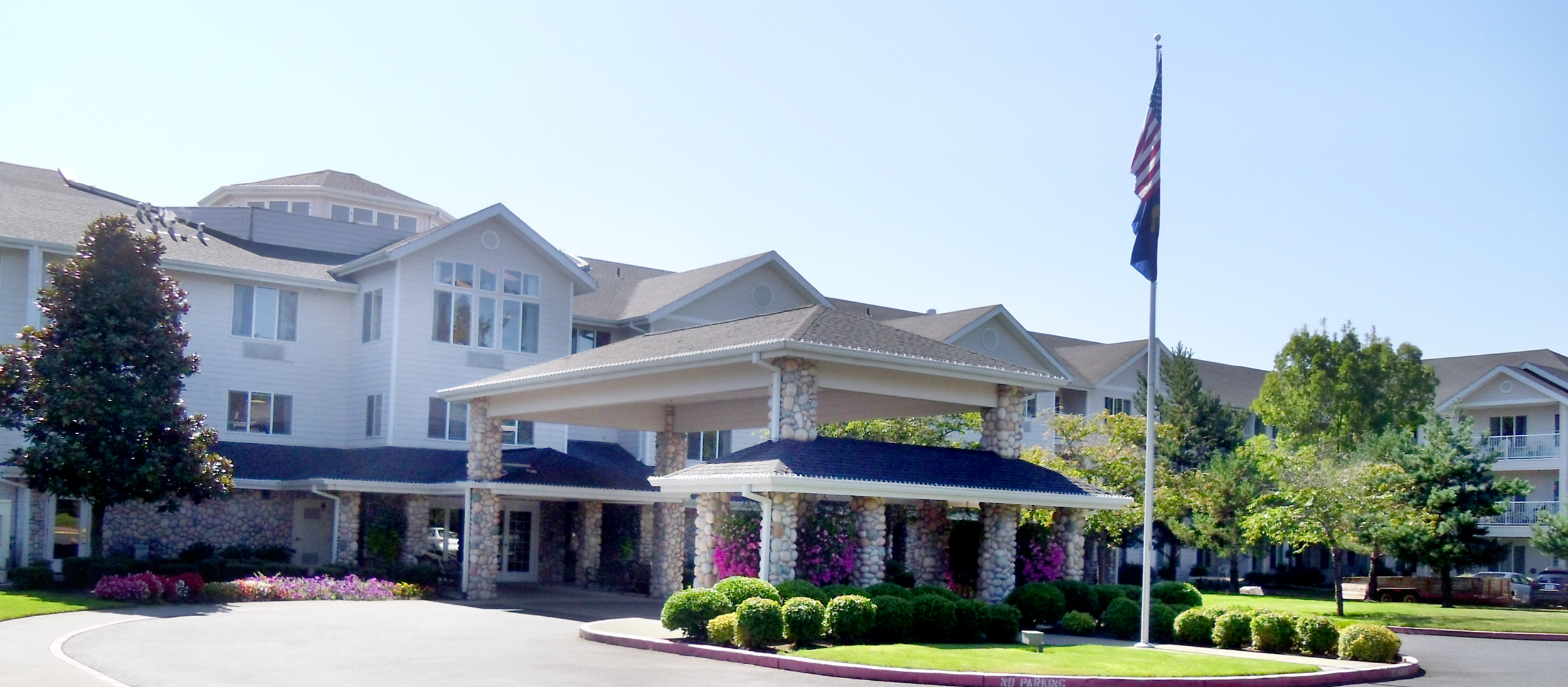 A large, three-story building with white exterior and stone columns under a porte-cochère. An American flag is on a flagpole in a grassy area with trimmed bushes. Flower beds are in front of the building and the sky is clear and blue.