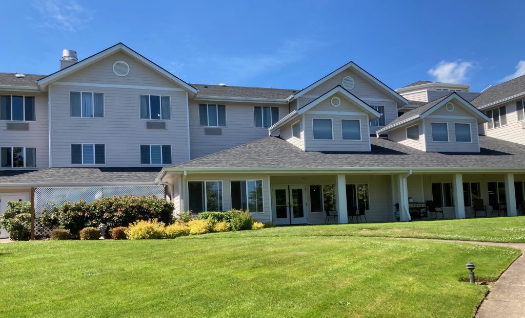 A large, light-colored three-story building with multiple gable roofs stands under a blue sky. The building has many windows and a covered patio area with seating. A well-maintained green lawn and bushes with flowers are in the foreground.