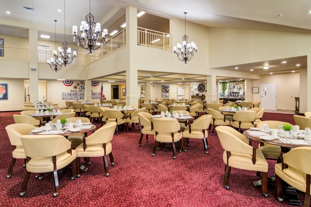 Spacious dining room with round tables set for meals, beige chairs, red carpet, and large chandeliers. Decor includes wall art and plants, and a mezzanine level overlooks the area. The atmosphere is bright and welcoming.