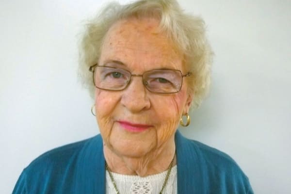 An elderly woman with short curly white hair, wearing glasses and gold hoop earrings. She is dressed in a blue cardigan over a white lace top and is smiling slightly, set against a plain white background.