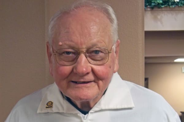 An older man with short white hair, wearing glasses and a white jacket, is smiling at the camera. The background features a beige wall and part of a ceiling with some greenery.