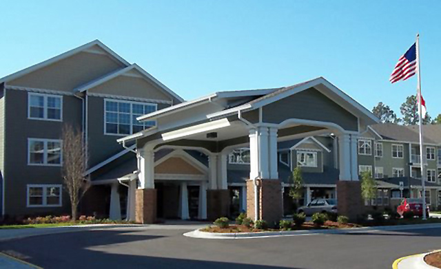 A modern, multi-story building with grey and white siding and brick accents. It features a covered entrance supported by large columns. An American flag is flying on a flagpole to the right, and landscaping with shrubs and trees surrounds the entrance.