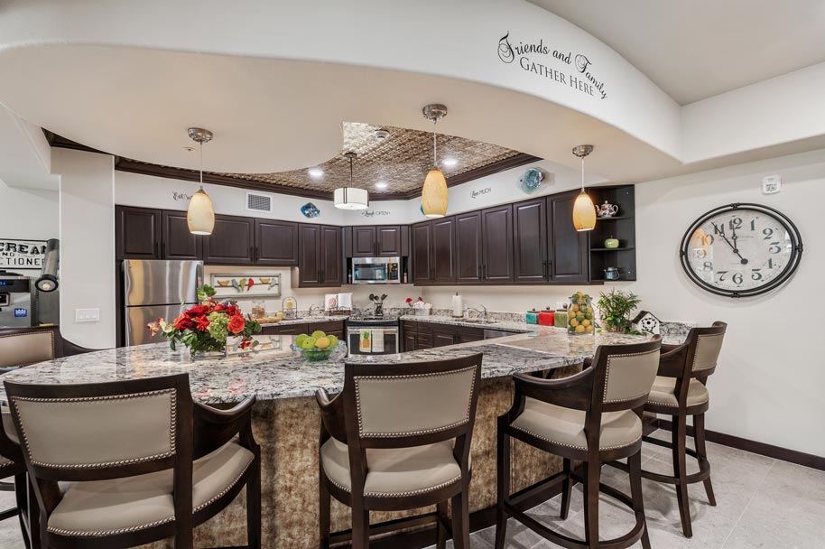 A spacious kitchen features a large granite island with six upholstered bar stools. The kitchen has dark wood cabinets, stainless steel appliances, and pendant lighting. A clock adorns the wall, and a decorative sign reads 