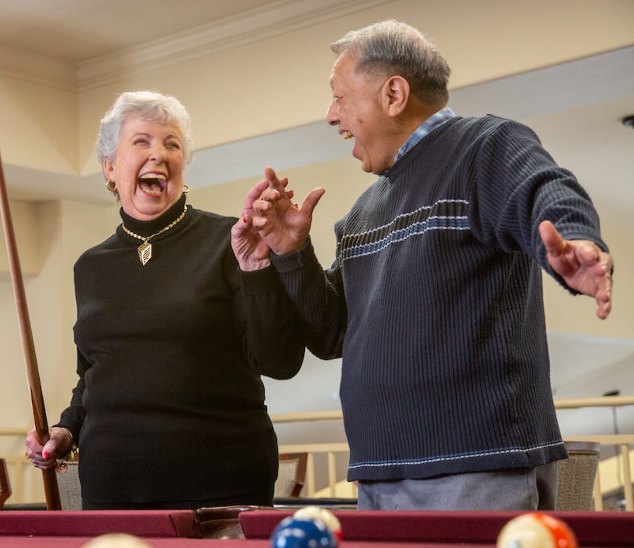 A senior woman holding a pool cue and a senior man standing next to her laughing joyfully. They are engaged in a game of pool, with colorful pool balls visible on the table in the foreground. Both are dressed casually and appear to be in a recreational setting.