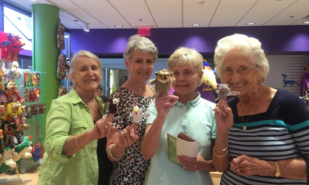 Four elderly women are smiling and posing inside a vibrant store filled with colorful items. Each woman is holding a finger puppet up. The store has purple walls and a variety of products on shelves in the background.