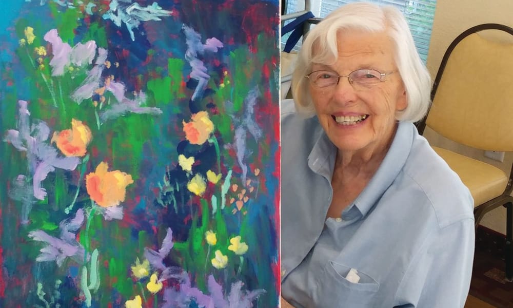 An elderly woman with white hair and glasses smiles while seated beside a colorful abstract floral painting. She is wearing a light blue shirt. The painting features vibrant yellows, purples, and greens, depicting lively flowers.