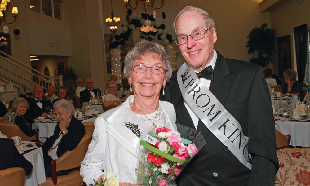 An elderly couple, crowned prom king and queen, smile warmly at the camera. The woman holds a bouquet of flowers and the man is dressed in a tuxedo. Both wear sashes indicating their titles. In the background, elegantly dressed seniors enjoy a festive event.