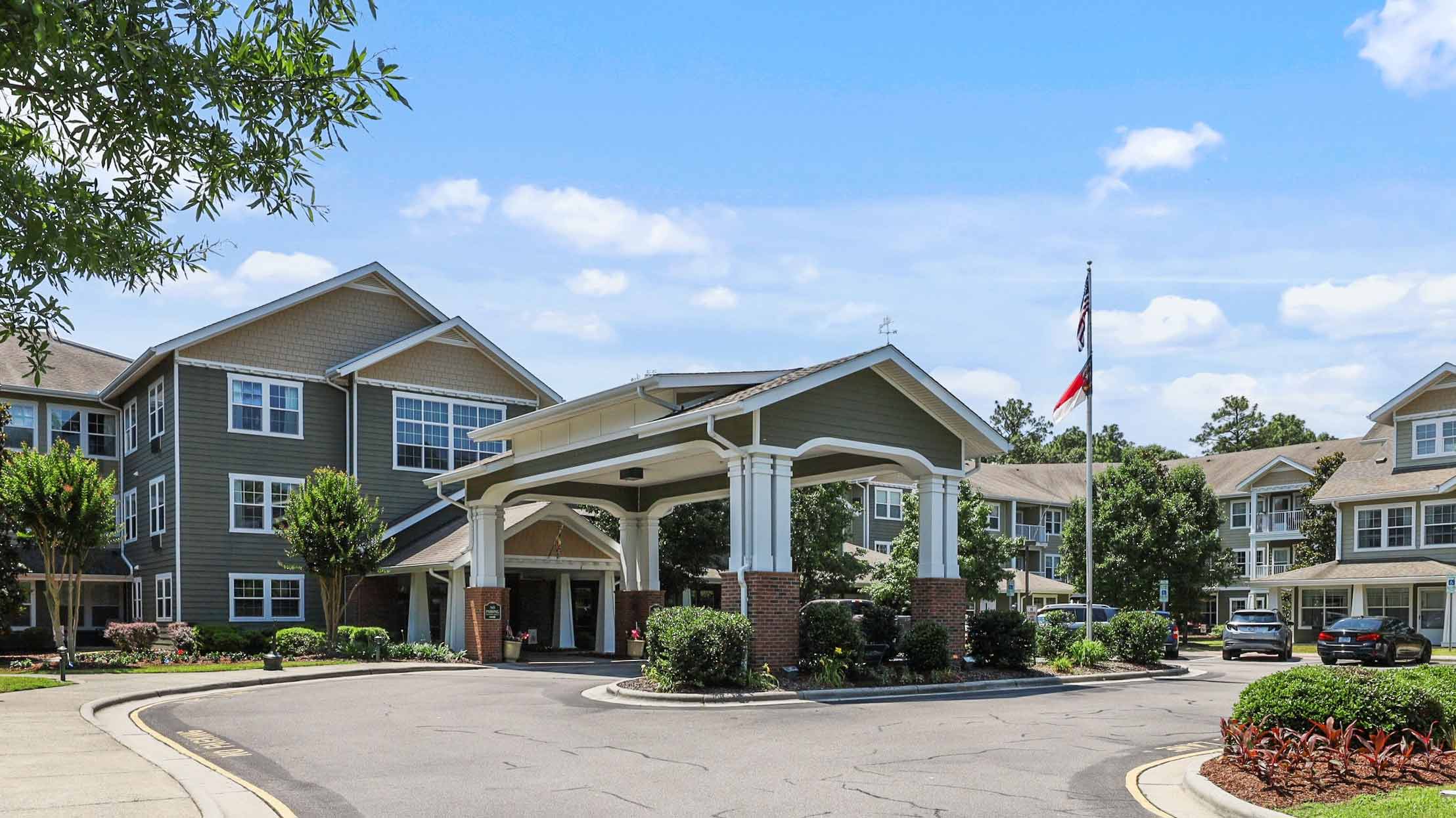 A large, two-story senior living facility with a covered entrance, surrounded by landscaping, trees, and parked cars. An American flag and another flag are flying on a pole in front. The sky is clear with a few clouds.