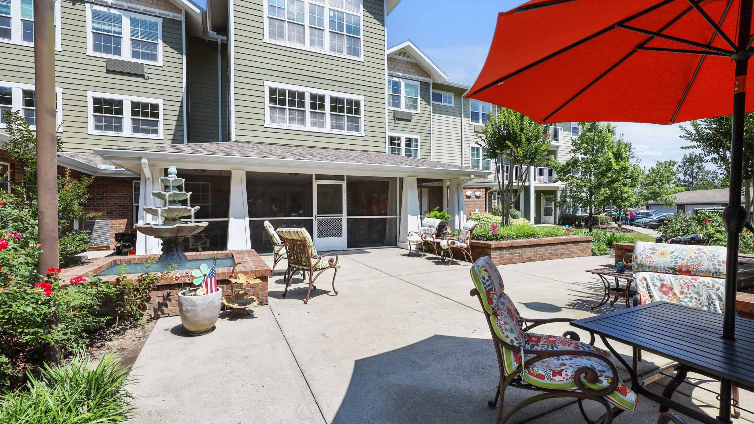 Sunny outdoor patio with colorful cushioned chairs, tables, a large red umbrella, and a decorative fountain surrounded by greenery in front of a multi-story residential building.