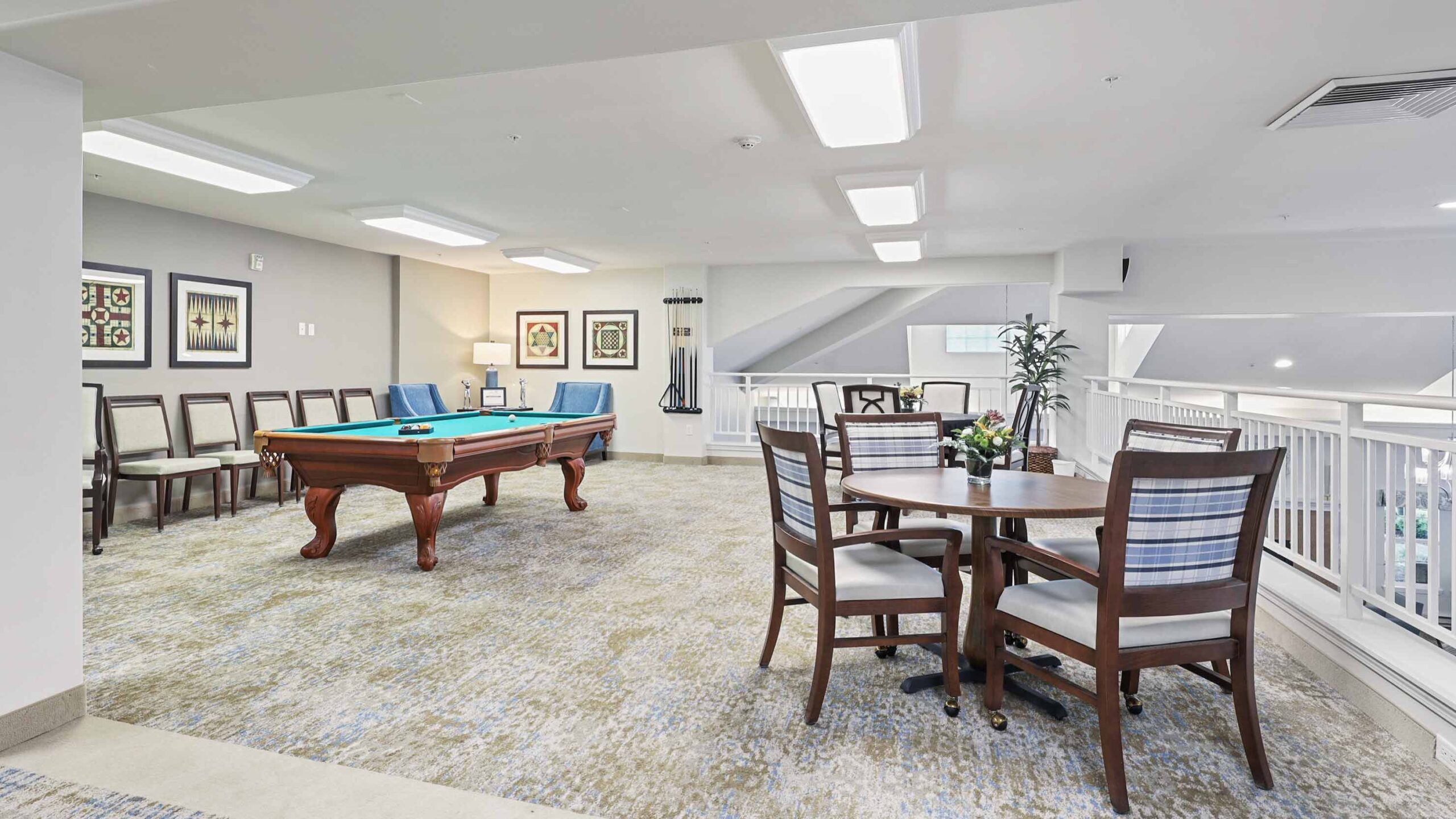 A bright recreation room with a pool table, cue sticks on the wall, framed artwork, a round table with four chairs, several lined-up chairs, and large windows letting in natural light.