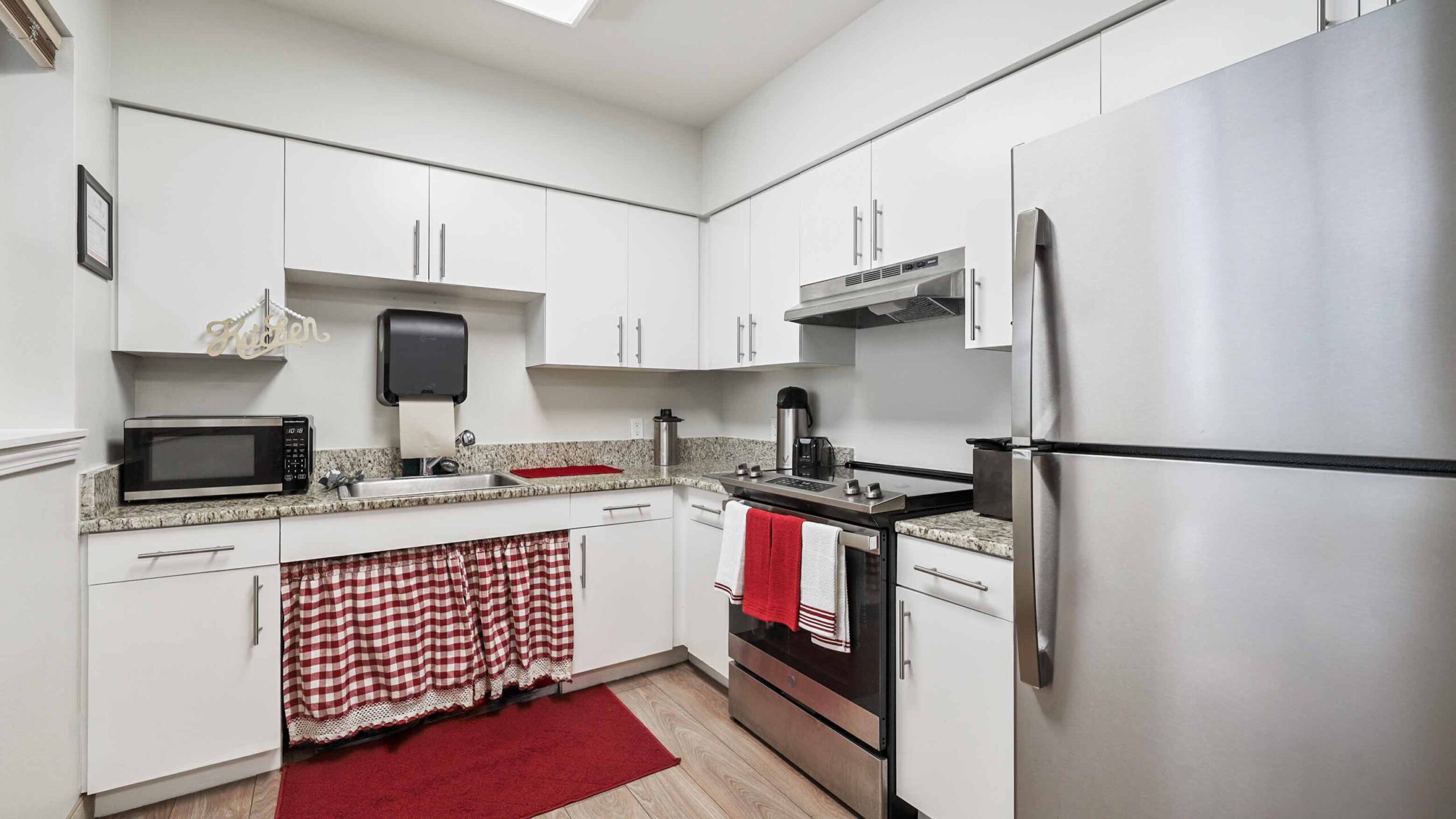 A modern kitchen with white cabinets, stainless steel appliances, a microwave, a red oven mitt and towels, and a red checkered skirt under the sink. There are red rugs on the wooden floor.