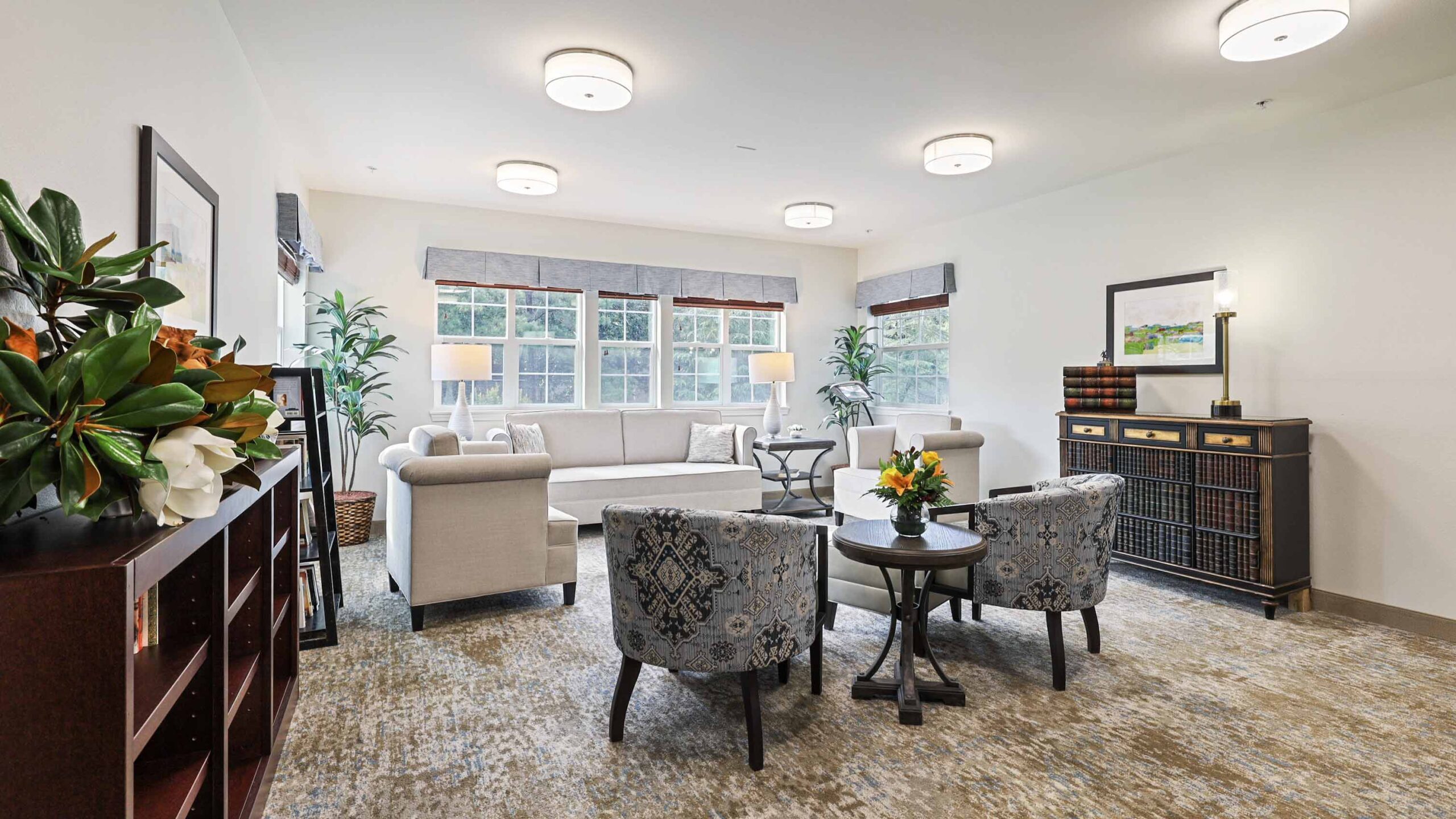 A bright, modern living room with large windows, cream-colored sofas, two patterned armchairs around a small table with flowers, a sideboard with books, and decorative plants.