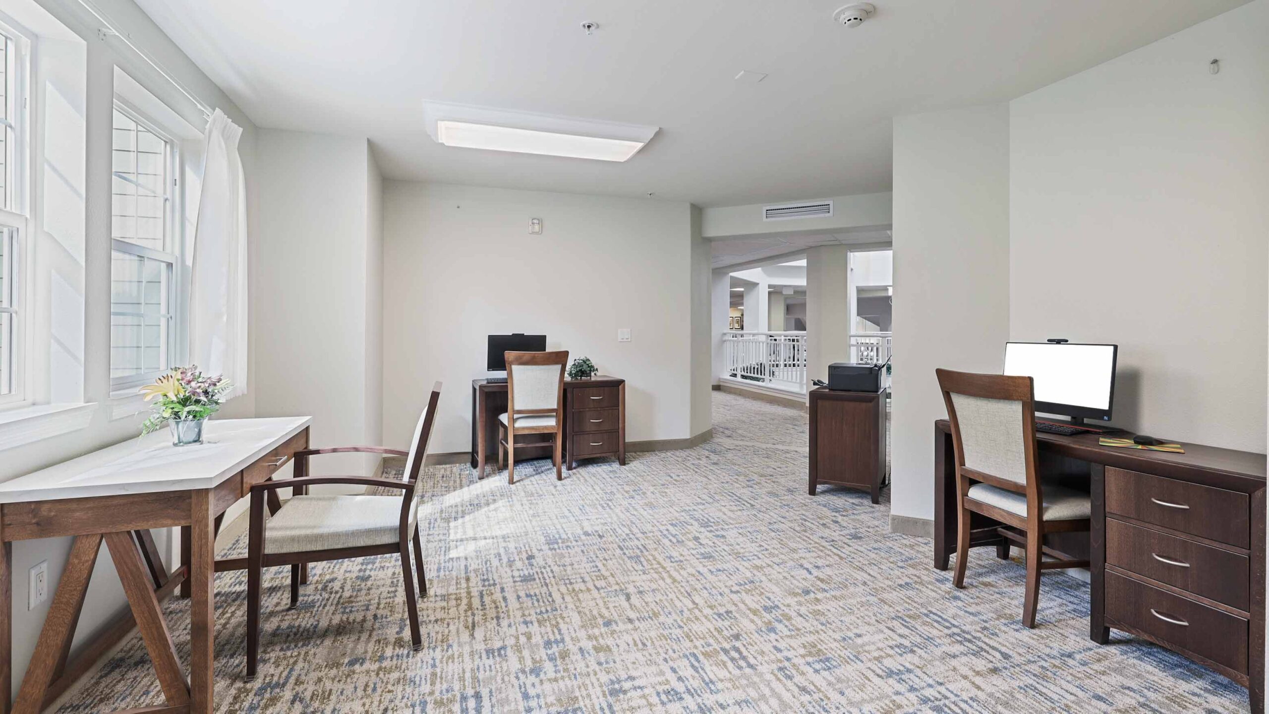 A bright office space with three wooden desks, each with a chair. Two desks have desktop computers, and one desk has a vase with flowers. The room has large windows and light-colored walls and carpet.