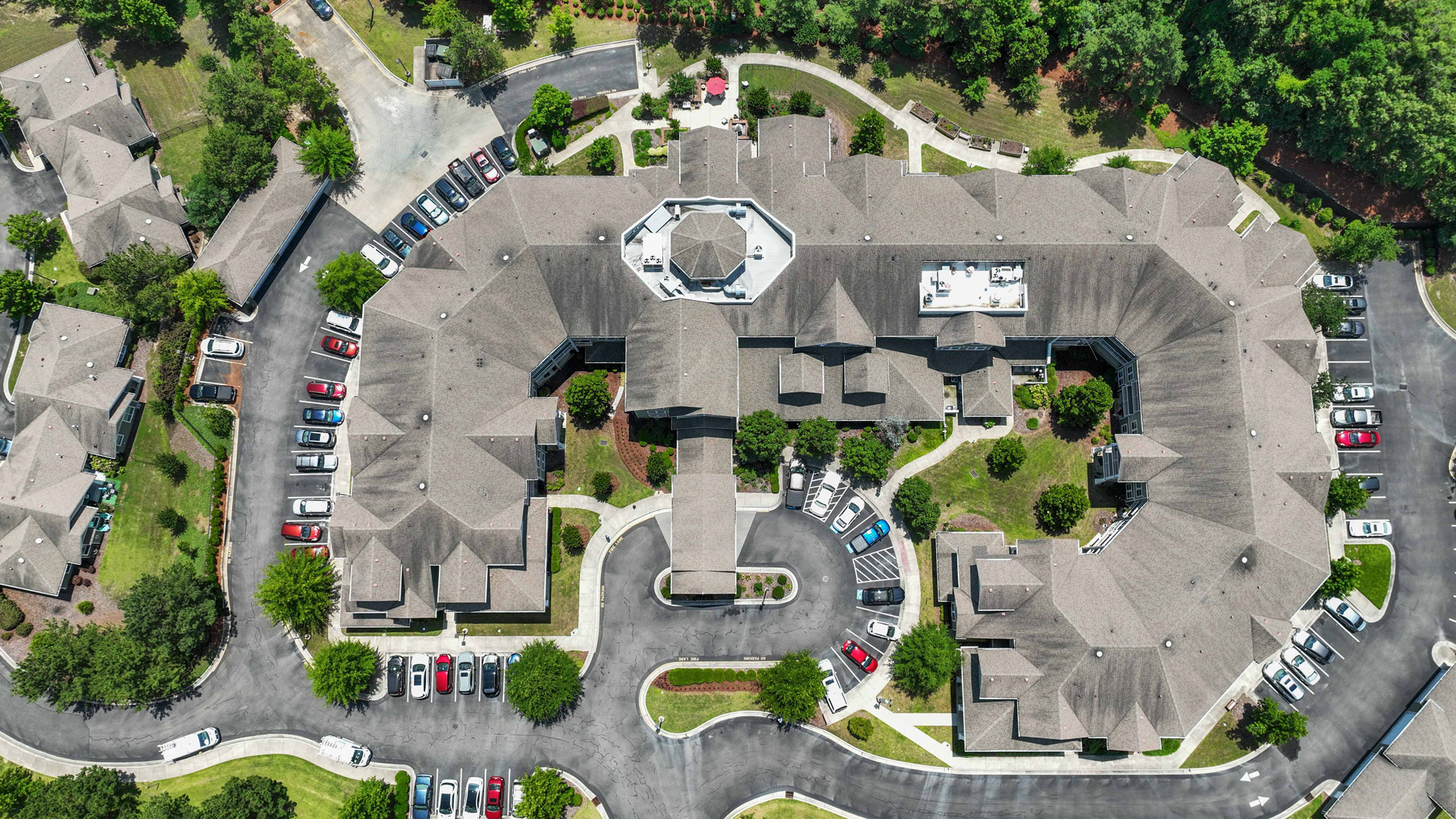 Aerial view of a large, multi-wing building with a central courtyard, surrounded by landscaped greenery and multiple parking lots filled with cars. Curved driveways and adjacent buildings are visible.