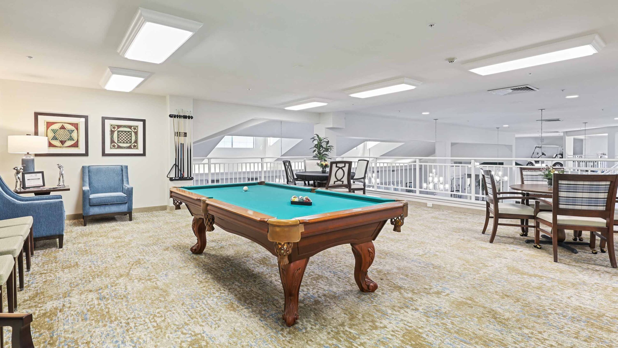 A bright, modern recreation room with a pool table in the center, surrounded by chairs, framed artwork, a potted plant, and a table with four chairs near a white railing overlooking the floor below.
