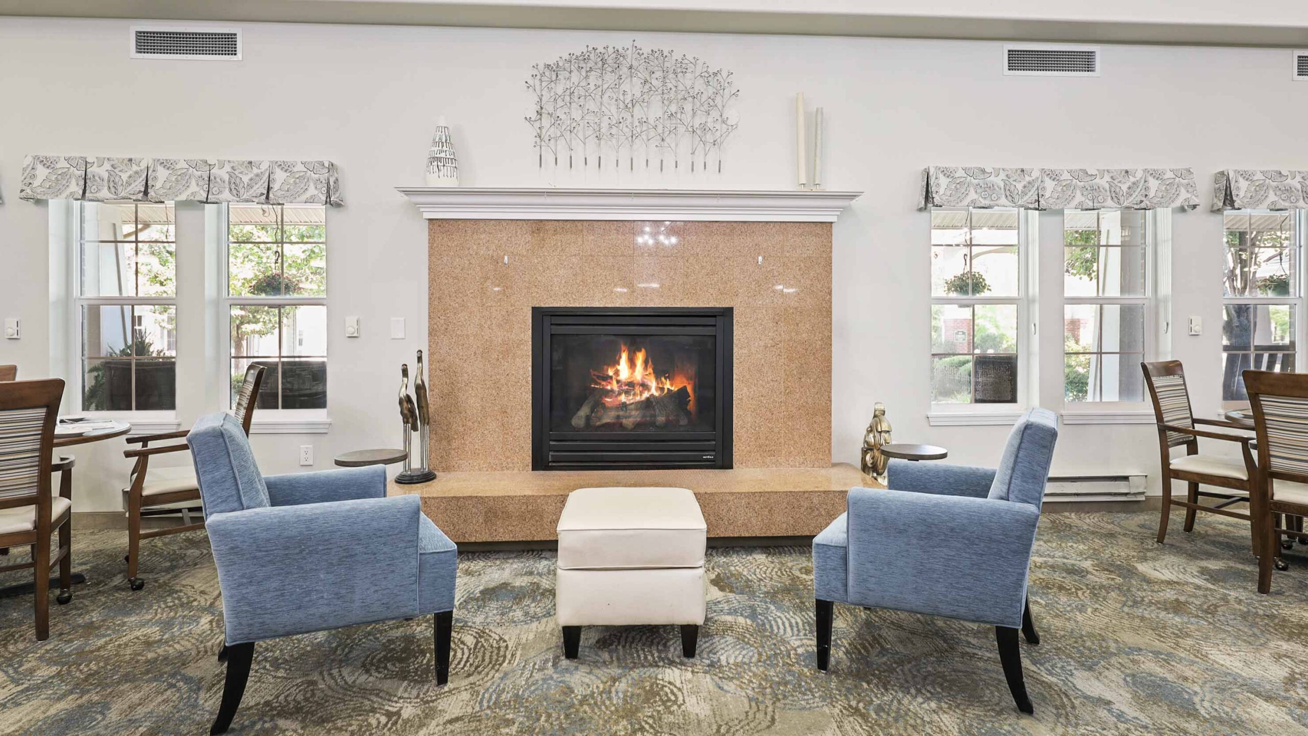 A cozy lounge with two blue armchairs and a white ottoman facing a fireplace. Large windows on each side let in natural light, and dining tables and chairs are seen in the background.