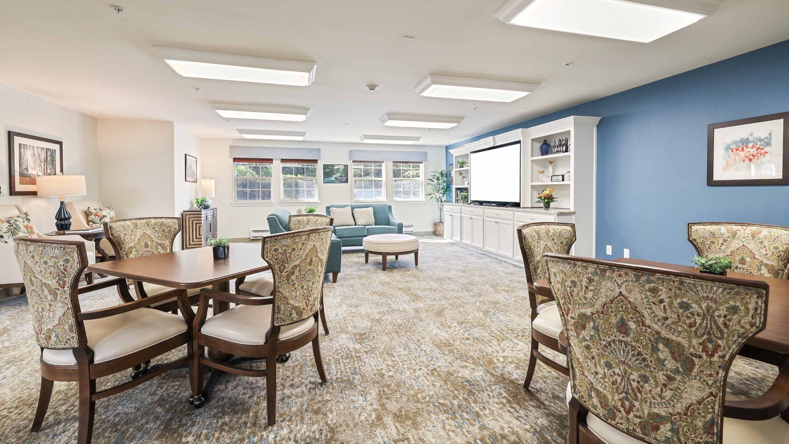 A bright, spacious lounge with patterned chairs around wooden tables, a soft carpet, a blue accent wall, large windows, and a seating area with couches and a TV entertainment center. Plants and artwork add a cozy touch.