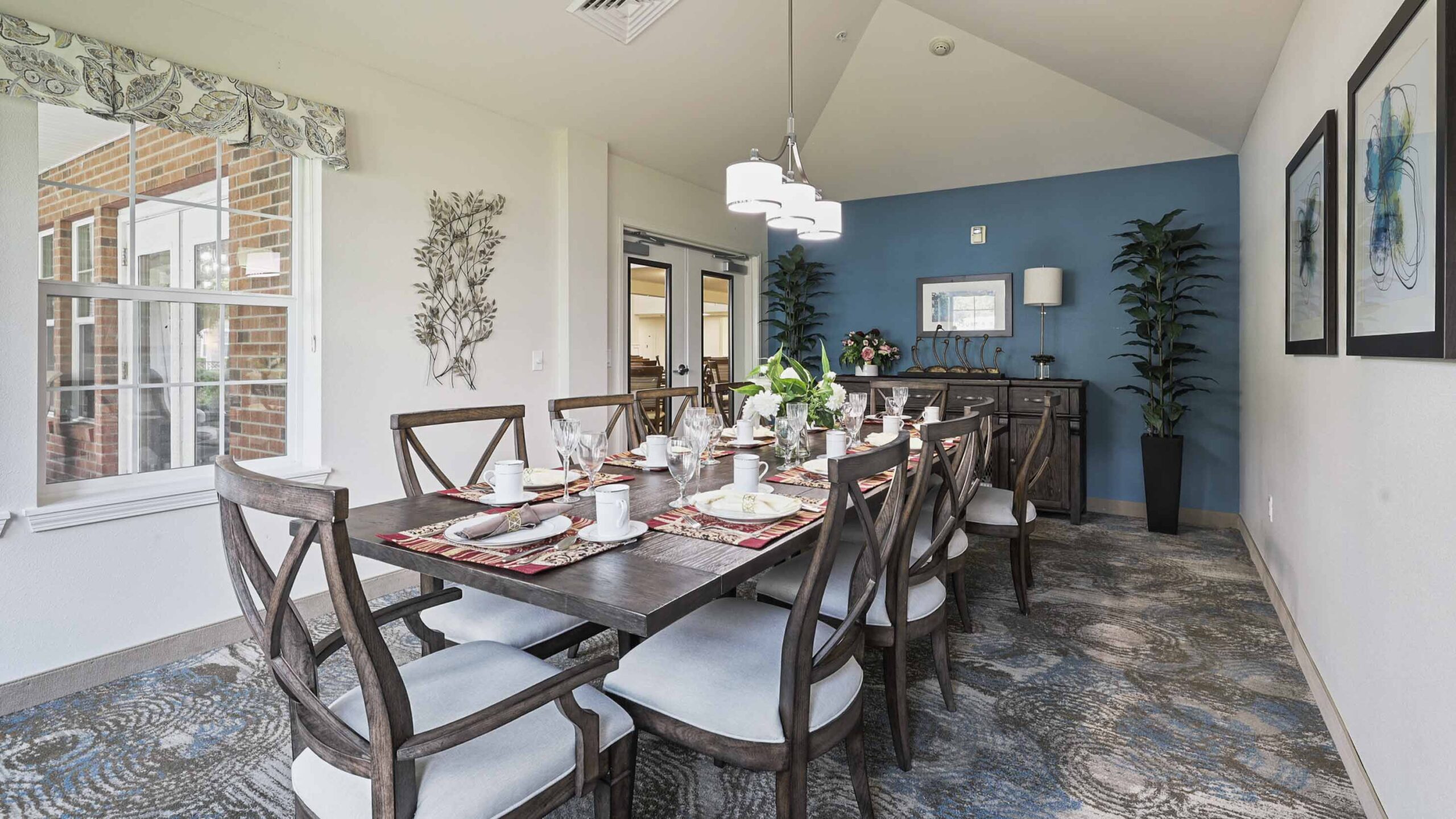 A modern dining room with a long wooden table set for eight, cushioned chairs, blue accent wall, decorative artwork, two tall plants, and a large window letting in natural light.