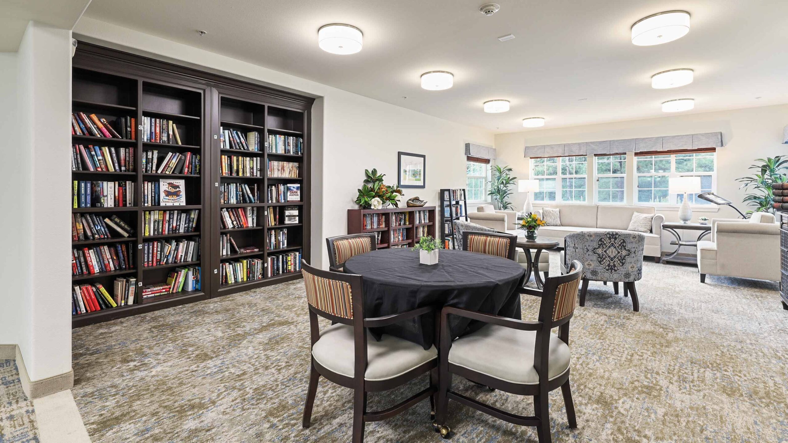 A cozy, well-lit lounge features a large bookshelf filled with books, a round table with four chairs, sofas, armchairs, potted plants, and large windows letting in natural light.