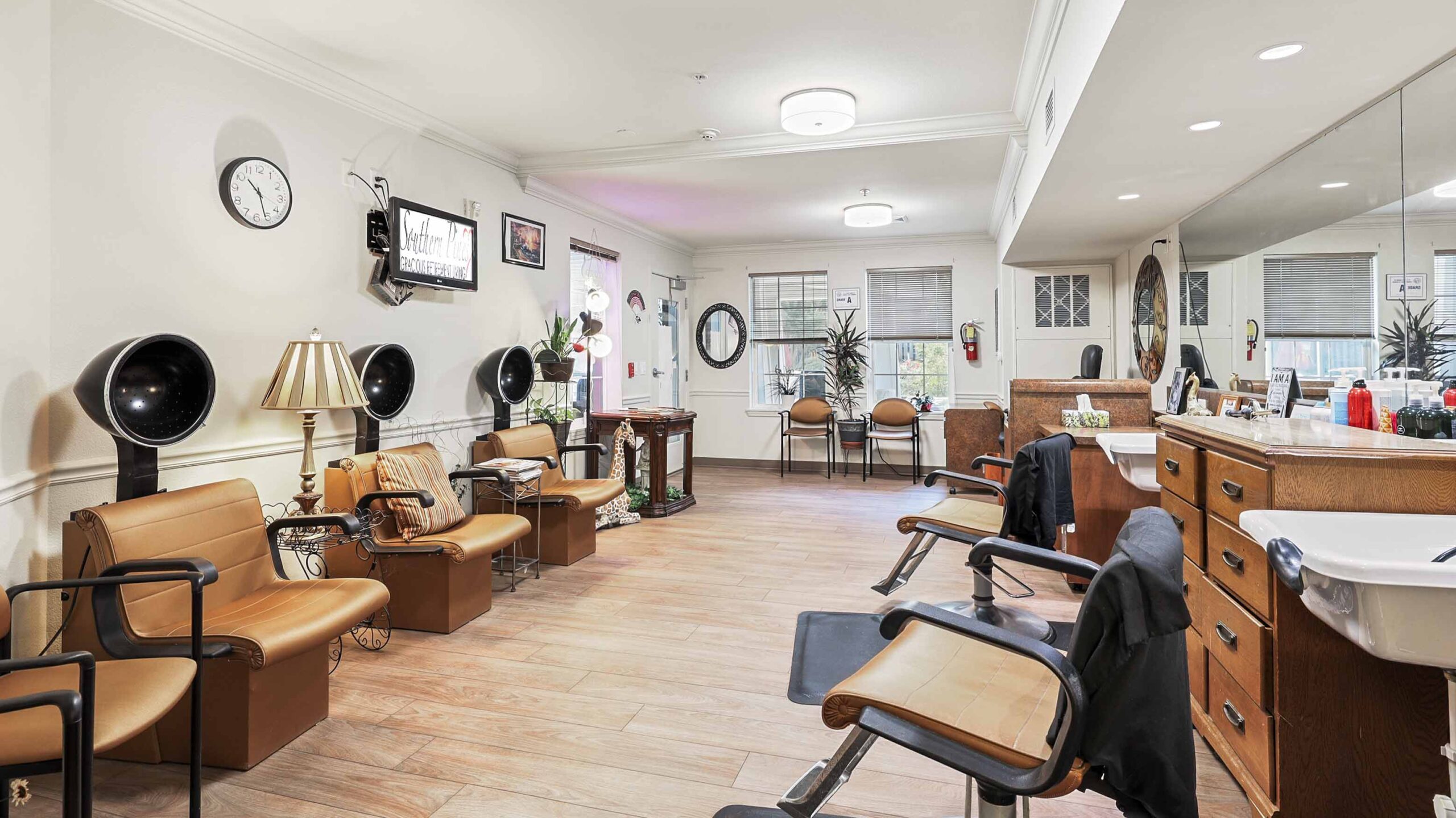 A spacious, vintage-style salon with tan chairs, retro hair dryers, wooden cabinets, large mirrors, and a bright, tidy atmosphere. The waiting area has armchairs and decor including lamps and wall clocks.