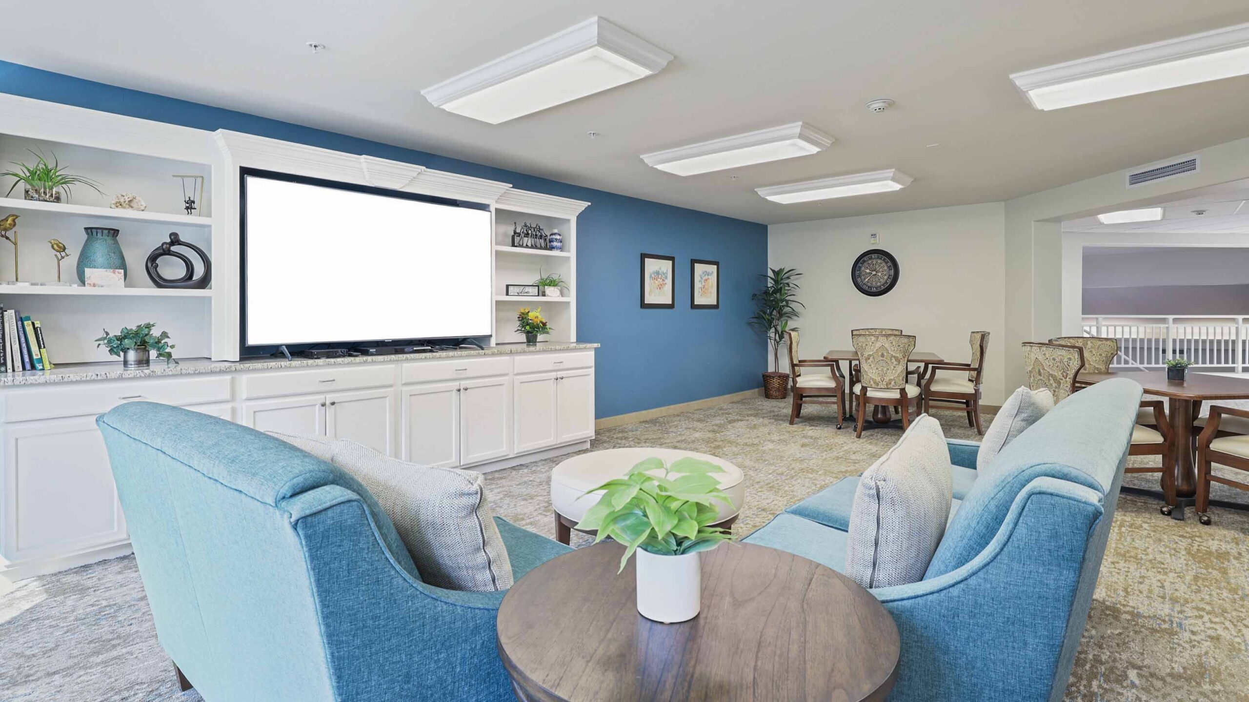 A bright lounge area features two blue armchairs, a small round table with a potted plant, a large white entertainment center with a blank TV screen, and dining tables in the background, all set on light carpeting.
