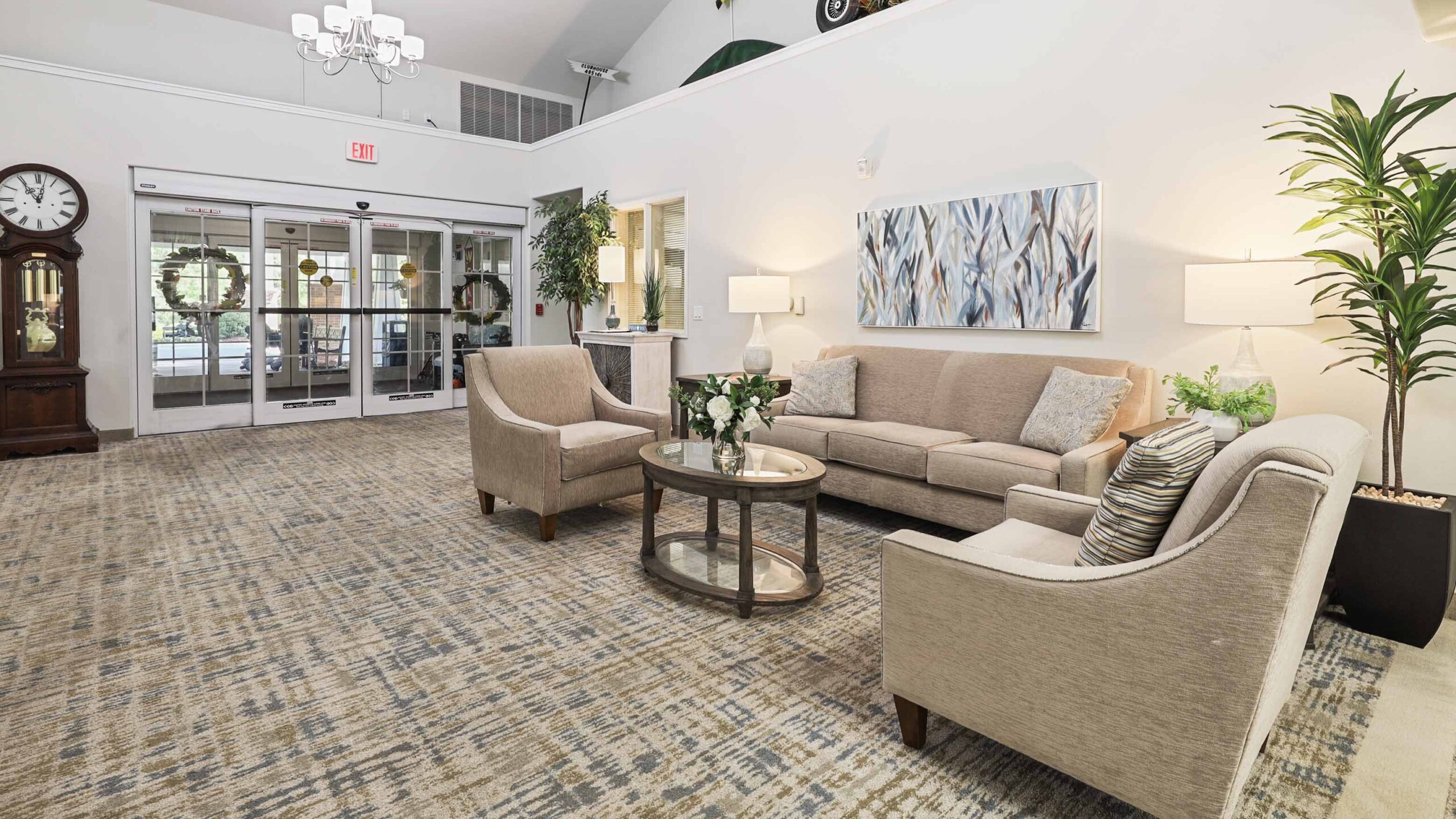 A bright, welcoming lobby with beige couches and armchairs around a glass coffee table, modern art on the wall, large plants, lamps, a patterned carpet, and glass double doors at the entrance.