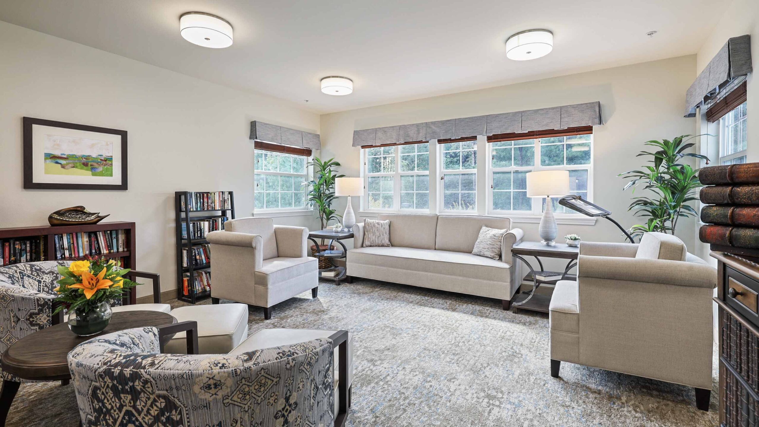 A bright, modern living room with large windows, neutral-toned sofas and armchairs, a patterned rug, bookshelves, potted plants, and a small round table with a vase of yellow flowers.