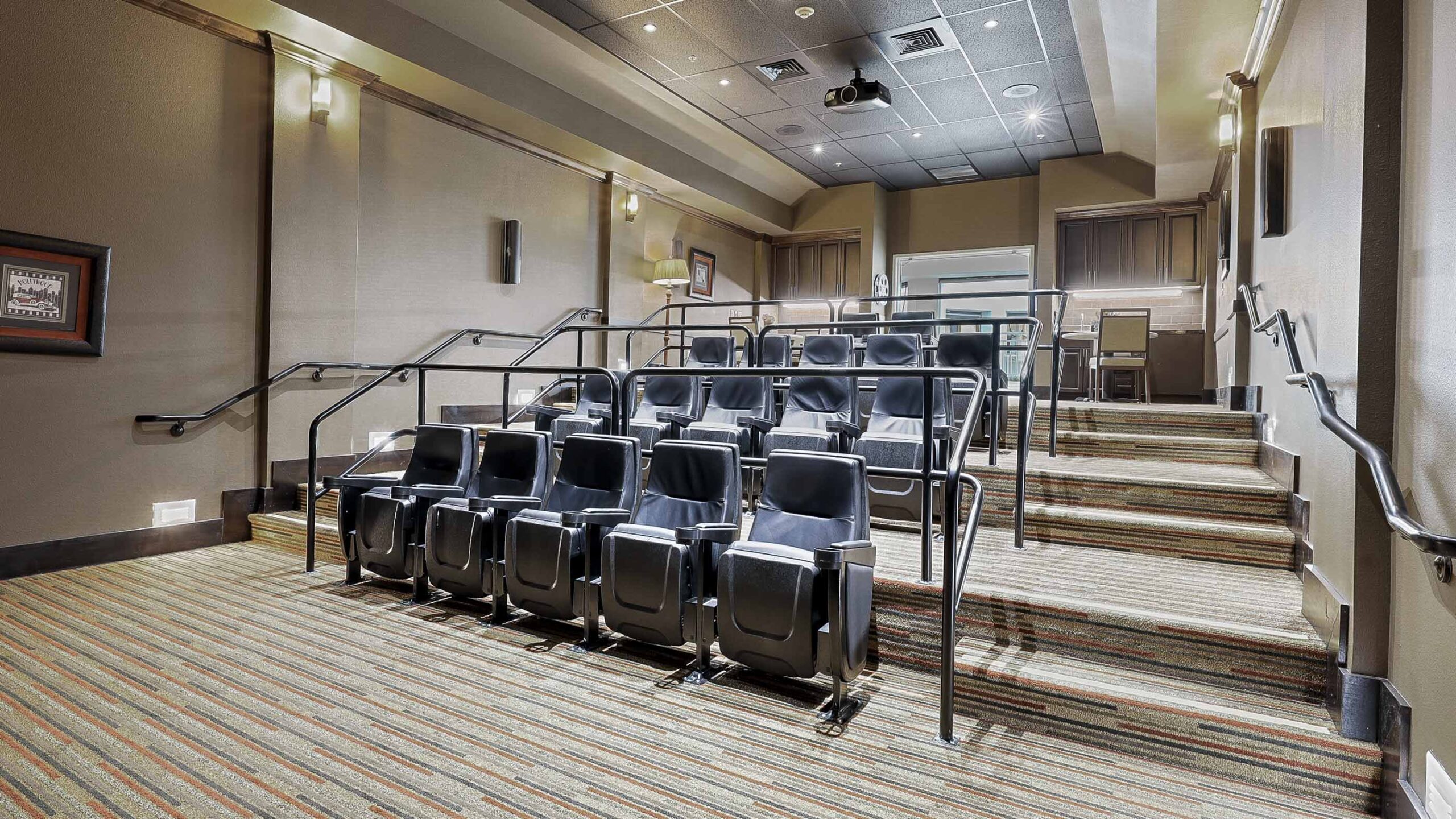 A small, modern home theater room with tiered seating, black cushioned chairs, carpeted floor, handrails, and soft wall lighting. The screen area is at the front with cabinetry in the back.