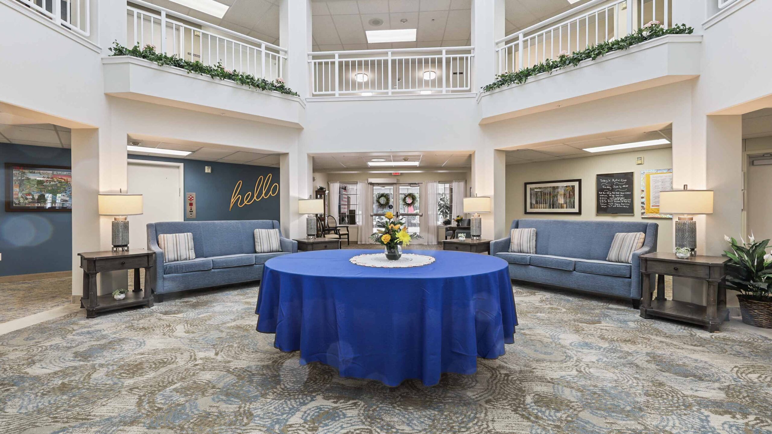 A spacious, bright lobby with a round table covered by a blue cloth and a flower centerpiece. Two blue couches with pillows face each other, and "hello" is written on the back wall. Decorative plants and lamps complete the cozy space.