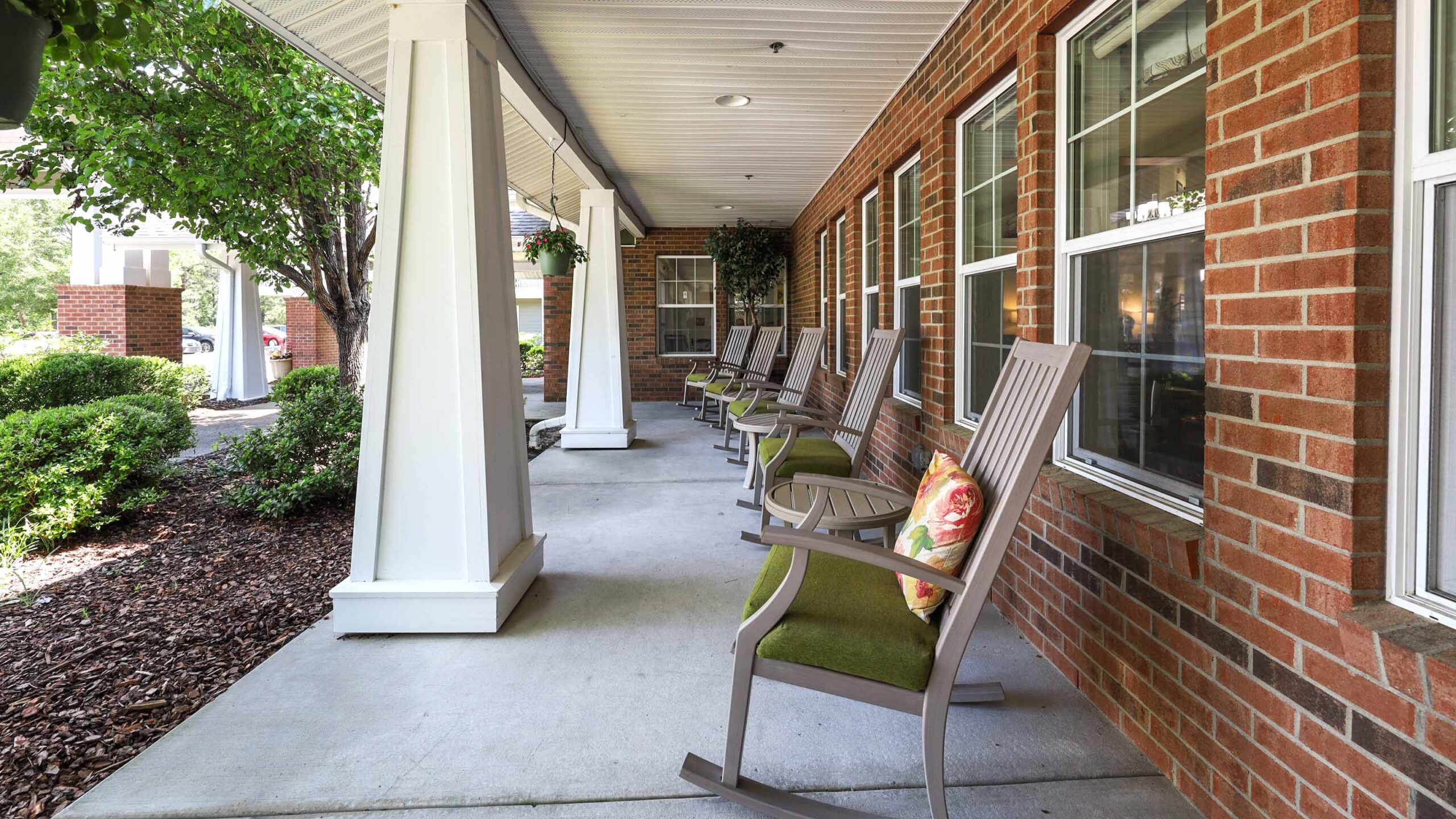 A covered brick porch with several wooden rocking chairs, some with cushions, overlooking a landscaped garden area with bushes and trees. Large white columns support the roof.
