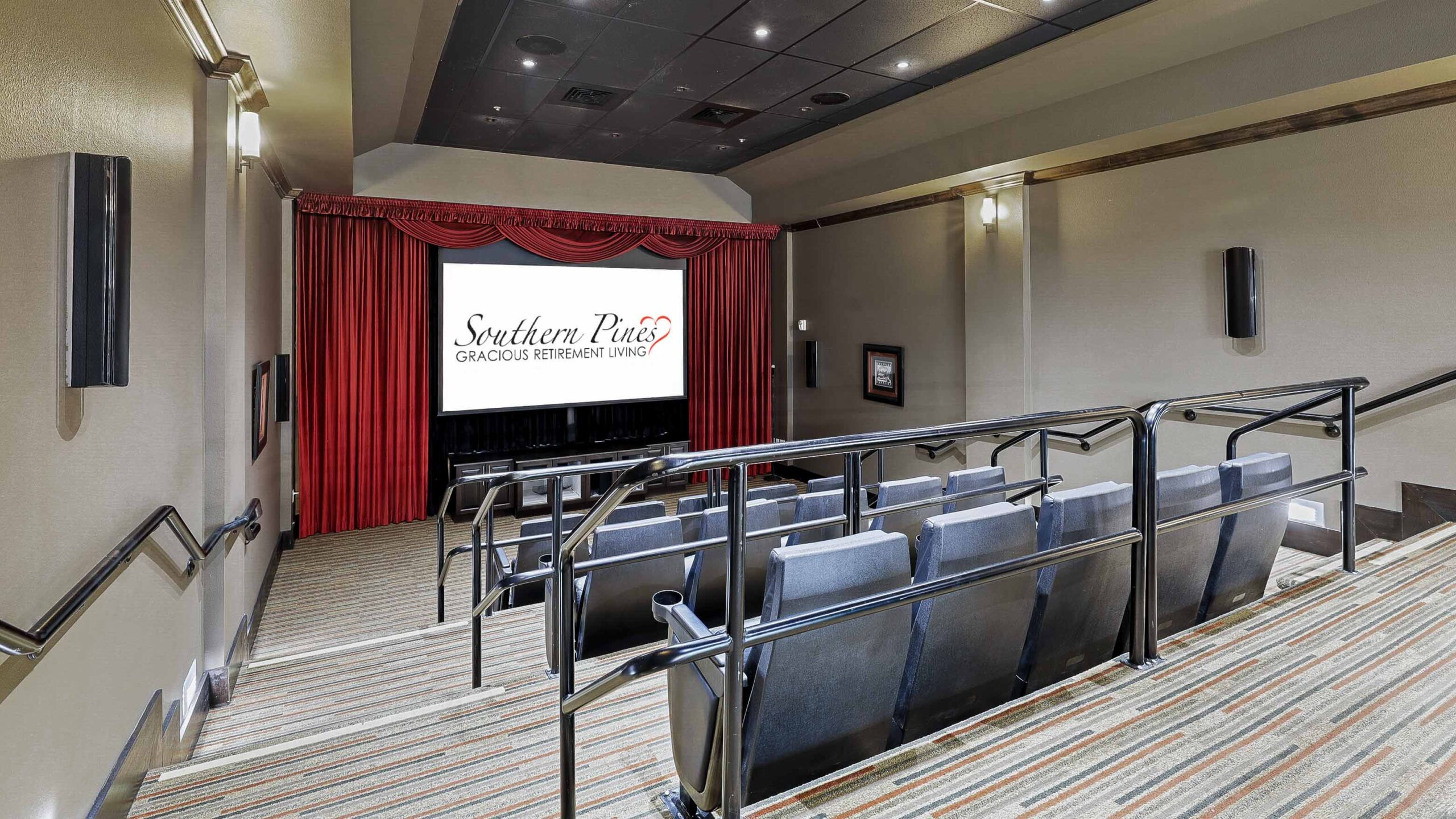 A small private theater with gray seats, a handrail-lined aisle, red curtains, and a large screen displaying “Southern Pines Gracious Retirement Living.” The room is softly lit with wall sconces.