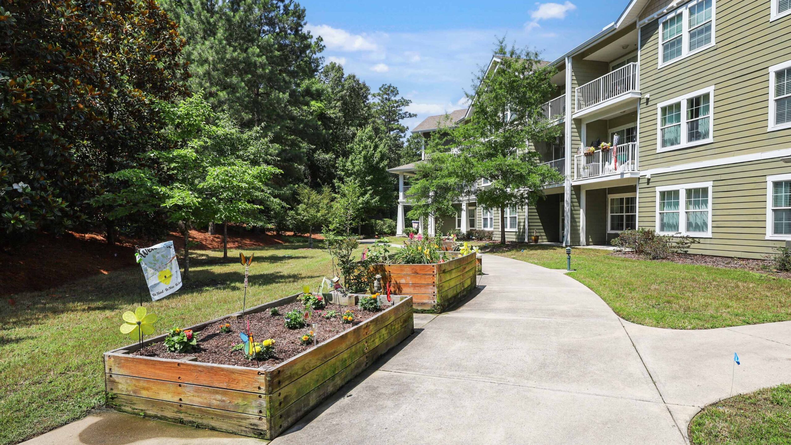 A concrete walkway curves past raised wooden garden beds filled with plants, in front of a green apartment building with balconies, surrounded by trees and grass on a sunny day.
