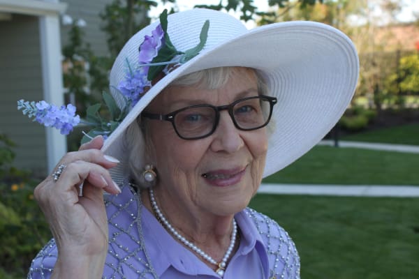 An elderly woman wearing glasses and a white hat with purple flowers on the brim smiles warmly. She wears a lavender blouse, pearl earrings, and a pearl necklace. A garden and building are visible in the background.