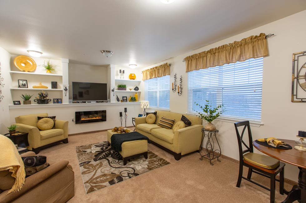 A cozy living room featuring light beige walls and carpet, two matching cream couches with yellow and green cushions, and a glass coffee table on a decorative rug. A TV and electric fireplace are built into a wall with shelves displaying decor. Large windows with tan valances let in natural light.