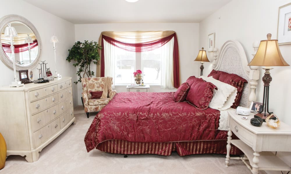A cozy bedroom featuring a bed with rich red bedding and fluffy pillows, a round mirror above a white dresser, an armchair with floral upholstery, and two nightstands with lamps. A large window, framed with red and beige drapes, illuminates the room.