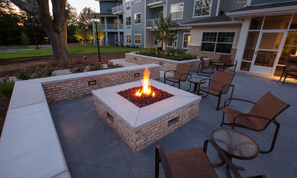 A cozy outdoor patio area with a central square fire pit surrounded by brick seating. The patio has several chairs and tables, and is adjacent to a modern building with large windows. Trees and a grassy lawn can be seen in the background.