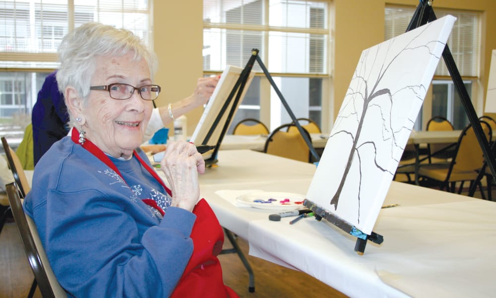 An elderly woman with glasses and short white hair is sitting at a table, holding a paintbrush and smiling. She wears a blue sweatshirt and a red apron. In front of her is a canvas with an outline of a tree. Other people painting can be seen in the background.