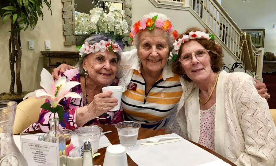 Three elderly women with flower garlands on their heads smile and pose together at a dining table. They are seated in a warmly lit room with a staircase in the background. One woman holds a cup, and the table is set with menus, flowers, and a pitcher of water.