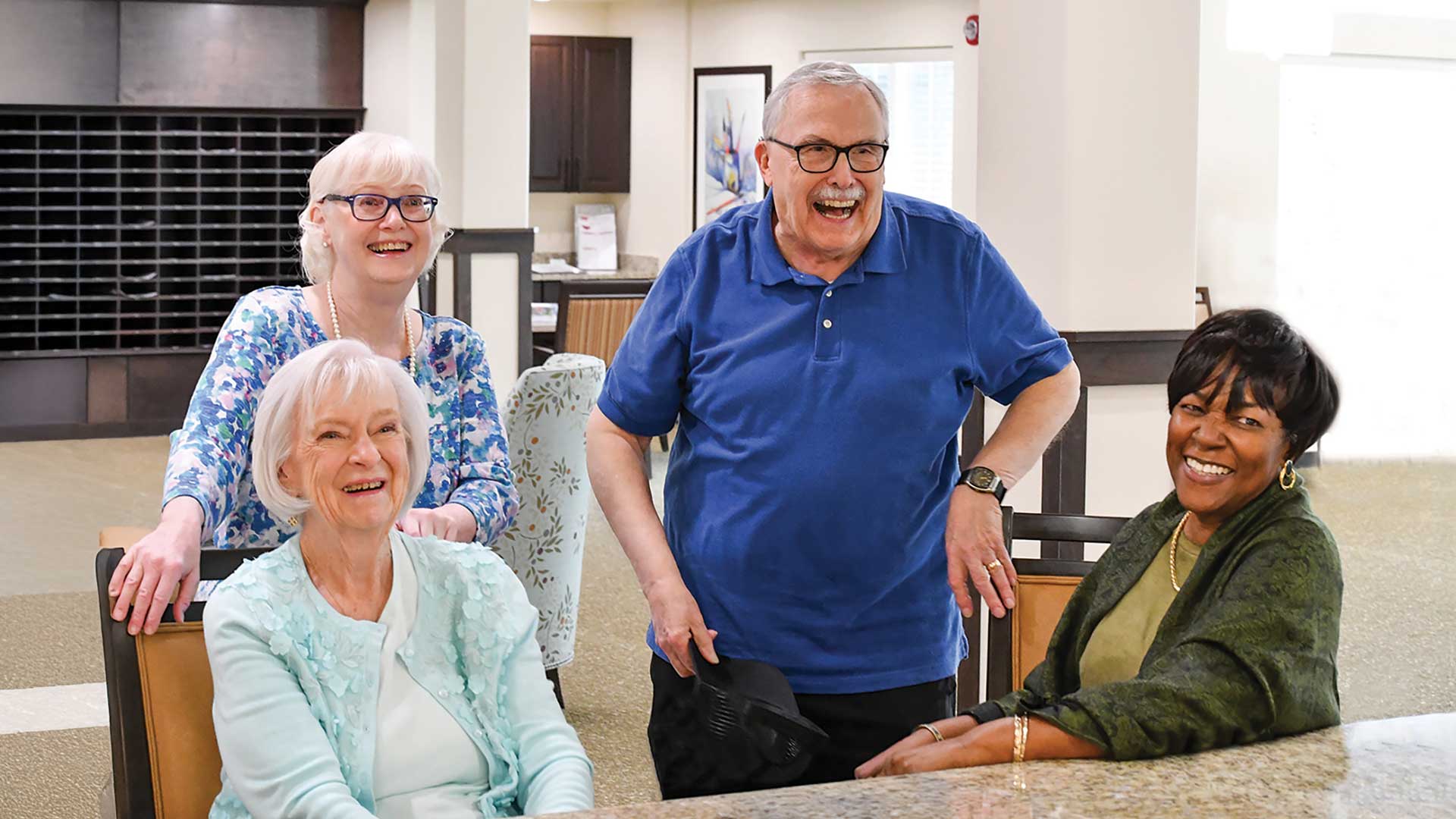 Four smiling older adults, three women and one man, sit and stand together around a table in a bright, modern communal space, appearing happy and engaged with each other.